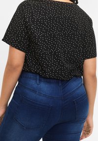 Schwarzes Hemd mit weißen Punkten, lockere Passform, kurze Ärmel. Kombiniert mit dunkelblauen Jeans mit Gesäßtaschen und glatter Textur.