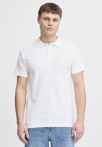 Hvit bomullspolo med flat krage, tre-knappers knappestolpe og ribbede ermer, kombinert med blå denimjeans.