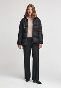 Czarna kurtka puchowa z kapturem, złotymi akcentami i detalem z logo; noszona na jasnoszarym swetrze, w połączeniu z czarnymi spodniami i botkami na obcasie.
