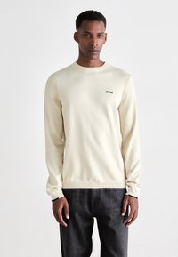 Suéter de manga larga crema claro con puños y dobladillo acanalados, que presenta un pequeño logo negro "BOSS" en el pecho. Llevado con jeans oscuros.