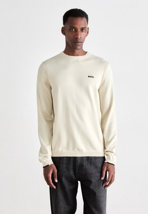 Light cream long-sleeve sweater with ribbed cuffs and hem, featuring a small black "BOSS" logo on the chest. Worn with dark jeans.