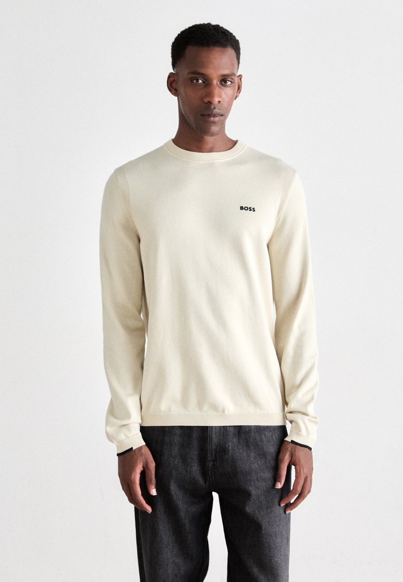 Suéter de manga larga crema claro con puños y dobladillo acanalados, que presenta un pequeño logo negro "BOSS" en el pecho. Llevado con jeans oscuros.