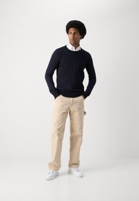 Navy sweater over a white collared shirt, paired with beige pants and white sneakers. The model stands upright, hands in pockets.