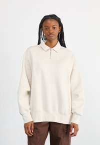 Ανοιχτό μπεζ φούτερ με μισό φερμουάρ στον γιακά, που διαθέτει ένα μικρό λευκό λογότυπο στο στήθος. Φτιαγμένο από μαλακό ύφασμα με άνετη εφαρμογή.