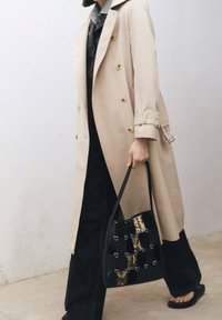 Trench-coat beige à double boutonnage, jean noir à jambes larges, et sac noir avec imprimé serpent et ferrures argentées. Sandales plates visibles.