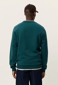 Τιρκουάζ φούτερ από βαμβακερό μείγμα, με στρογγυλή λαιμόκοψη, ριμπ μανσέτες και τελείωμα. Φορεμένο πάνω από τζιν, από πίσω. Συνοδεύεται από σκούρο καπέλο.