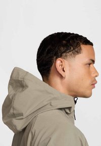 Ljusbeige huva jacka med slät textur, utrustad med dragsko och sydda detaljer, modellerad från en sidoprofil.