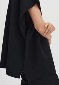 Haut noir ample au design drapé, en tissu texturé, avec un détail de bouton sur le côté. Il possède des manches larges et une coupe ouverte.