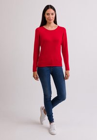 Roter Pullover mit langen Ärmeln aus weichem Stoff, runder Ausschnitt, kombiniert mit blauen Skinny-Jeans und weißen Sneakers, steht vor einem schlichten Hintergrund.