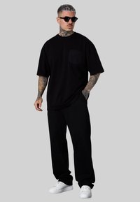 T-shirt en coton noir avec une poche sur le devant, associé à un pantalon de survêtement noir et des baskets blanches, offrant une coupe décontractée et une texture douce.