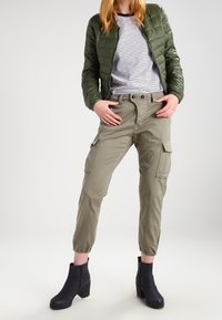 Olijfgroene gewatteerde jas over een wit met marineblauw gestreept shirt, gecombineerd met lichtkhaki cargobroek en marineblauwe enkellaarsjes.