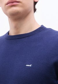 Άνδρας που φοράει μπλε ναυτικό πουλόβερ με στρογγυλή λαιμόκοψη και μικρό λευκό λογό τυπο Levi's στο στήθος, εμφανίζεται από το λαιμό έως το πάνω μέρος του κορμού.