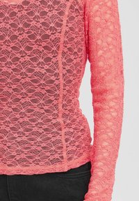 Top a maniche lunghe in pizzo corallo con motivo floreale, tessuto trasparente e orlo arrotondato. Abbinato a jeans neri in vita.