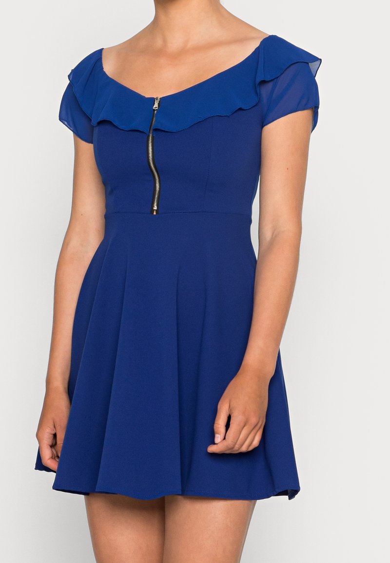 Robe courte bleue avec encolure bardot à volants, manches courtes transparentes, fermeture éclair avant et jupe évasée portée par une personne.