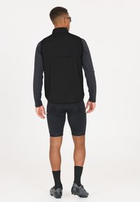 Svart väst med hög krage, bärs över en långärmad topp, kombineras med svarta shorts och svarta träningssko. Enkelt design, inga synliga logotyper.