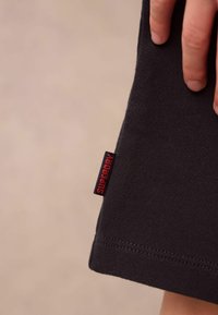 Černá textilní látka s hladkým povrchem, na lemu vyznačená červenou nálepkou "SUPERDRY". Detailní záběr na ruku, která drží okraj.