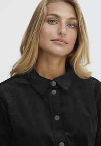 Chemise noire à boutons avec col, dotée de cinq boutons métalliques, faite d'un tissu texturé, présentant un design simple et épuré.