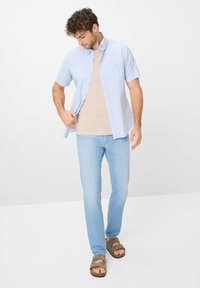 Camicia a maniche corte, a quadri blu chiaro sopra una maglietta beige, indossata con jeans blu chiaro e sandali marroni. Vestibilità casual con dettagli ai bottoni.