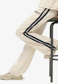 Crème-kleurige broek met zwarte zijstrepen en de tekst "ATHLETICS" boven de rechterknie. Zachte stof, recht gesneden design, gedragen met lichte sneakers.