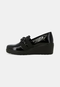 Scarpa wedge in vernice nera con cinturino elastico e dettaglio decorativo in catena nera sul lato, su sfondo grigio chiaro.