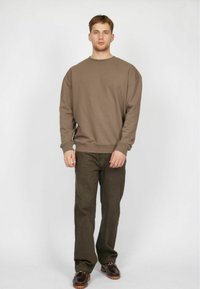 Sudadera marrón de corte relajado y cuello redondo, combinada con pantalones verde oscuro y zapatos marrones. Diseño sencillo, sin patrones.