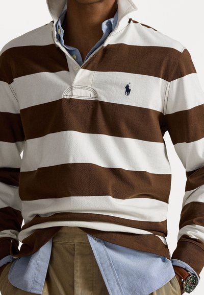 Tricou de rugby în dungi maro și alb cu guler, având un mic logo albastru. Purtați peste o cămașă deschisă la culoare. Material din bumbac.