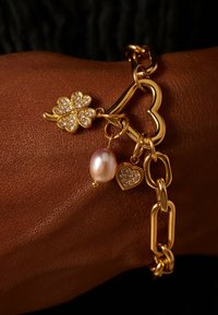 Gouden schakelarmband met een vierbladige klaver bedel met rhinestones, een parelmoer kraal en een andere hartbedel met rhinestones.
