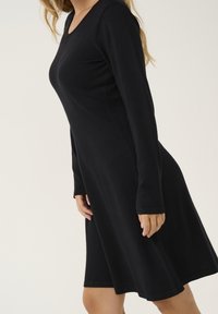 Robe noire à manches longues avec un col en V, une silhouette évasée en ligne A et une texture douce. Présente des accents cousus à l'ourlet et aux manches.