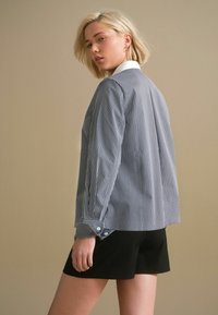 Šedobílý pruhovaný blazer s bílým límcem, knoflíkovými manžetami a volným střihem, doplněný černými šortkami. Hladká textura.