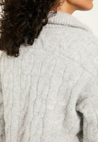 Grauer Pullover mit Kabelstrickmuster, geripptem Kragen und lockerer Passform. Das Material wirkt weich und gemütlich.