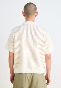 Short-sleeve, cream-colored knit polo with openwork pattern. Features a ribbed collar and hem, accentuated by a relaxed fit and subtle texture.