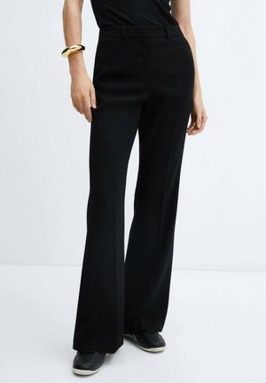 Femme portant un pantalon noir taille haute à jambes larges, un haut noir, des baskets noires avec semelles blanches et un bracelet doré massif au poignet gauche.
