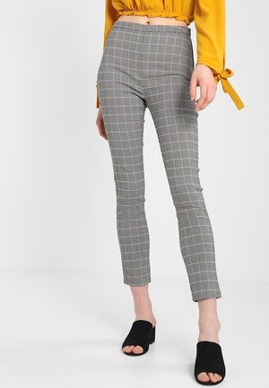 Kvinde iført tætsiddende grå ternede bukser, en cropped sennepsgul bluse og sorte åbne hælede mules, stående foran en hvid baggrund.