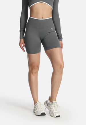 Kvinde iført grå højttaljede cykelshorts og kort langærmet top med hvide kanter, kombineret med hvide sportssko, stående på hvid baggrund.