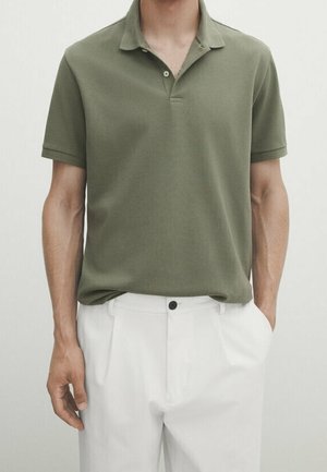 Mann trägt ein olivgrünes kurzärmeliges Polohemd, das in weiße plissierte Hosen mit einem schwarzen Knopf gesteckt ist.