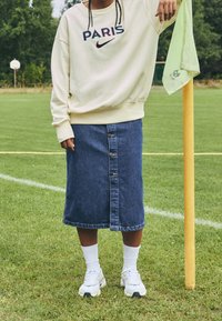 Persona vestida con un sudadera color crema con el texto "PARIS", una falda vaquera azul de botones, calcetines blancos y zapatillas deportivas, de pie junto a un banderín de esquina en un campo deportivo amarillo.
