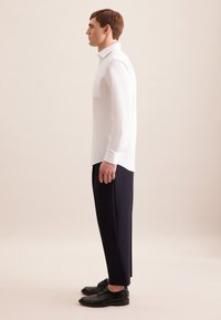 Camicia bianca a maniche lunghe con taschino, abbinata a pantaloni blu scuro. Modello che indossa scarpe eleganti nere, mostrato di profilo su uno sfondo neutro.