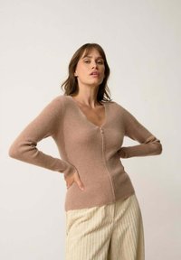 Cárdigan de punto color marrón claro con mangas largas, escote redondeado y cremallera frontal. Textura suave con sutil acanalado vertical.