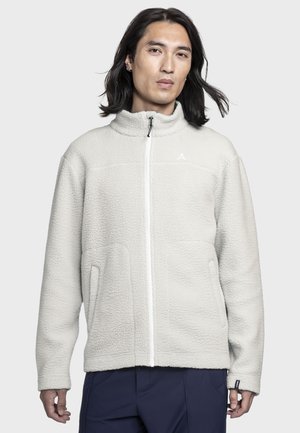 Mann mit schulterlangem schwarzen Haar, trägt eine cremefarbene Fleecejacke mit Reißverschluss und dunkelblaue Hose, steht vor einem einfarbigen Hintergrund.