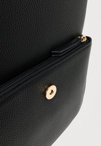 Μαύρη τσάντα από δέρμα με υφή, με κλείσιμο με χρυσό κουμπί και διαμέρισμα με φερμουάρ. Η επιφάνεια έχει πέτρινη υφή για επιπλέον λεπτομέρεια.