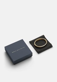 Kuldne käevõru musta nahast kotikese peal, sinise karbi kõrval, mille peal on kuldne "Daniel Wellington" tekst. Lihtne, tsilindriline disain.