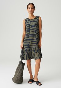 Sleeveless dress in dark green with abstract navy pattern. Features a flared hem and soft texture. Paired with a woven black bag and black slides.