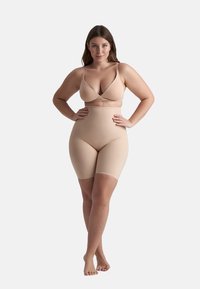Donna in piedi con le mani sui fianchi, indossa pantaloni shapewear a vita alta color nude e un reggiseno abbinato, su sfondo neutro.