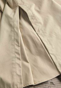 Close-up van beige stof met gedetailleerd stiksel en plooien over een bruine en zwarte geruite textiel eronder.