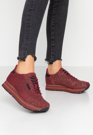 Fødder iført røde glitrende sneakers med sorte såler og brune striber, kombineret med sorte skinny jeans med frynset kant på hvidt gulv.