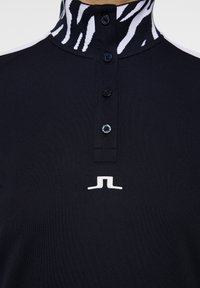 Svart polo med ett zebra-mönstrat krage, har knappstängning och en vit logotyp på framsidan. Slät textur och atletisk passform.