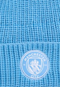 Bonnet tricoté bleu clair avec texture côtelée. Présente un logo circulaire de Manchester City en bleu clair et blanc à l'avant.