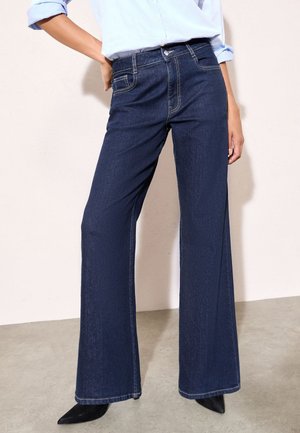 Person iført mørkeblå vidde jeans, lyseblå opkrogede skjorte og sorte spidse sko, der står på en lys gulv mod en bleg væg.