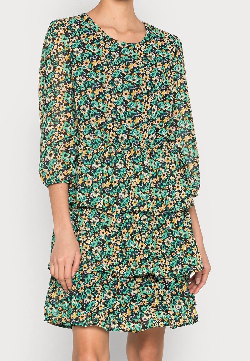 Femme portant une robe à fleurs avec des fleurs vertes, jaunes et blanches sur fond noir, dotée de manches trois-quarts et d'une jupe à volants.