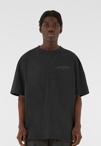 Sort oversized bomulds t-shirt med korte ærmer, der har et lille lilla logo på venstre brystområde. Blødt, let struktureret stof.
